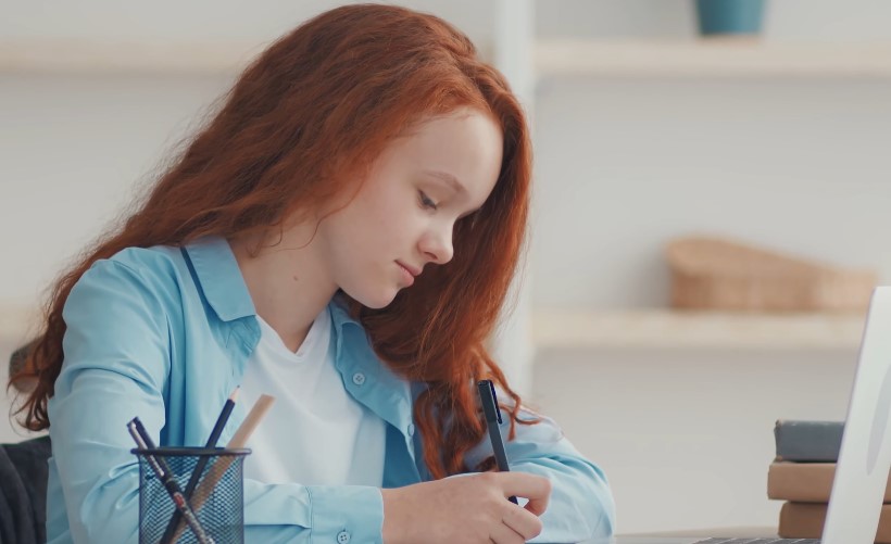 A girl with red hair is writing in a notebook, focused on her task