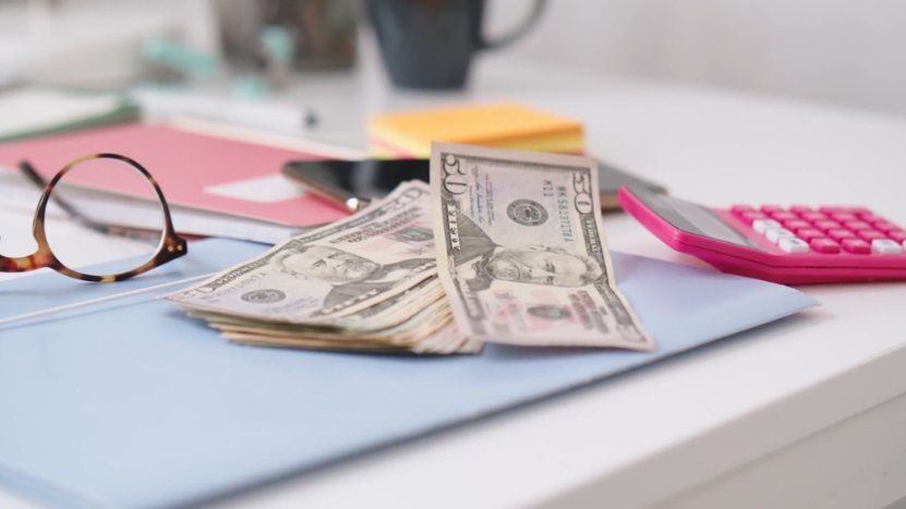 A stack of dollar bills sits on a desk beside notes and a calculator