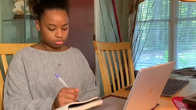 A Young Woman Is Taking Notes While Working on Her Laptop at A Dining Table, Balancing Work and Studies