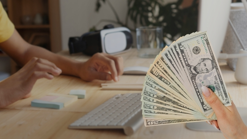 A Person Works at A Desk While a Hand Holds a Stack of Cash, Questioning if Online Learning Is Truly More Affordable