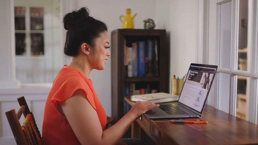 A Woman in An Orange Top Is Working on Her Laptop, Researching Online University Programs at A Desk in A Home Office