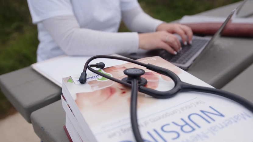 A Healthcare Worker Typing on A Laptop with A Stethoscope and Nursing Book on The Table