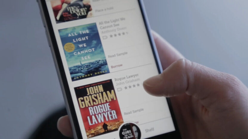 Close-up of a person browsing e-books on a smartphone, featuring book covers including 'All the Light We Cannot See' and 'Rogue Lawyer'.