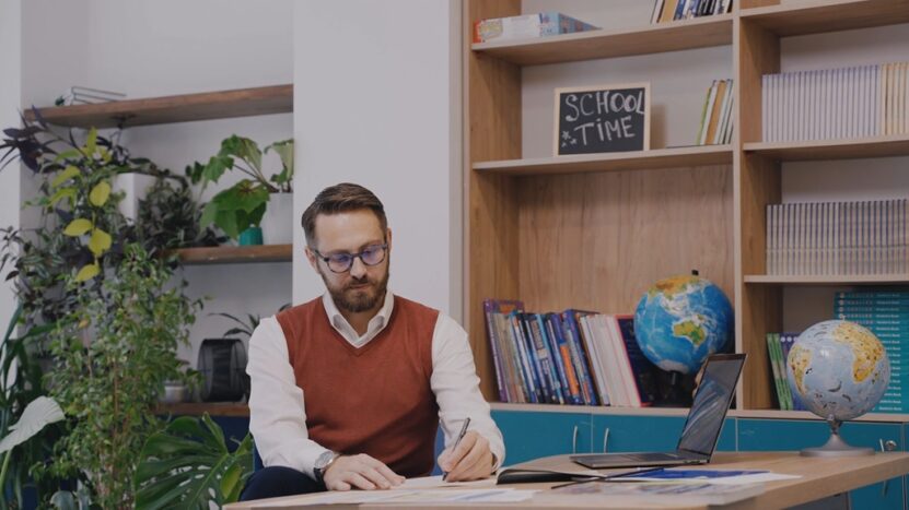 A man sits at a desk in a classroom filled with globes and bookshelves