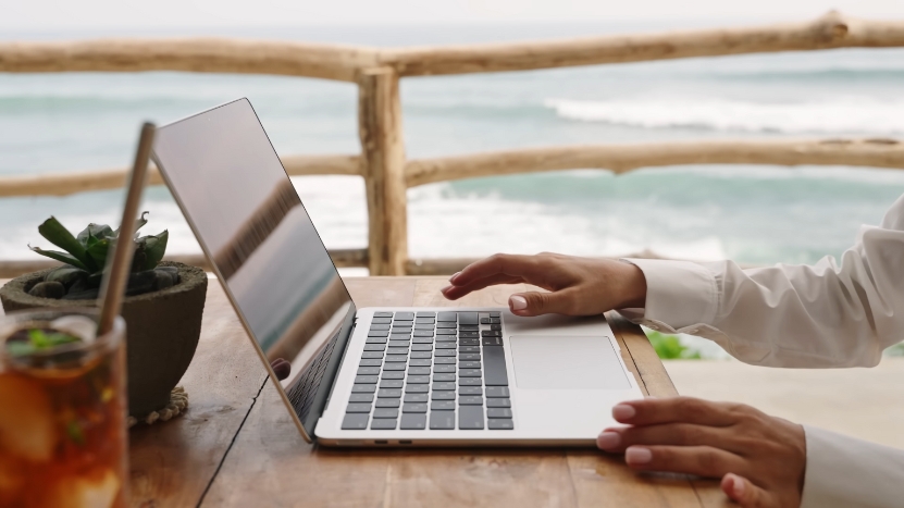 Person Working on A Laptop at A Wooden Table with An Ocean View, a Potted Plant, and A Drink Nearby